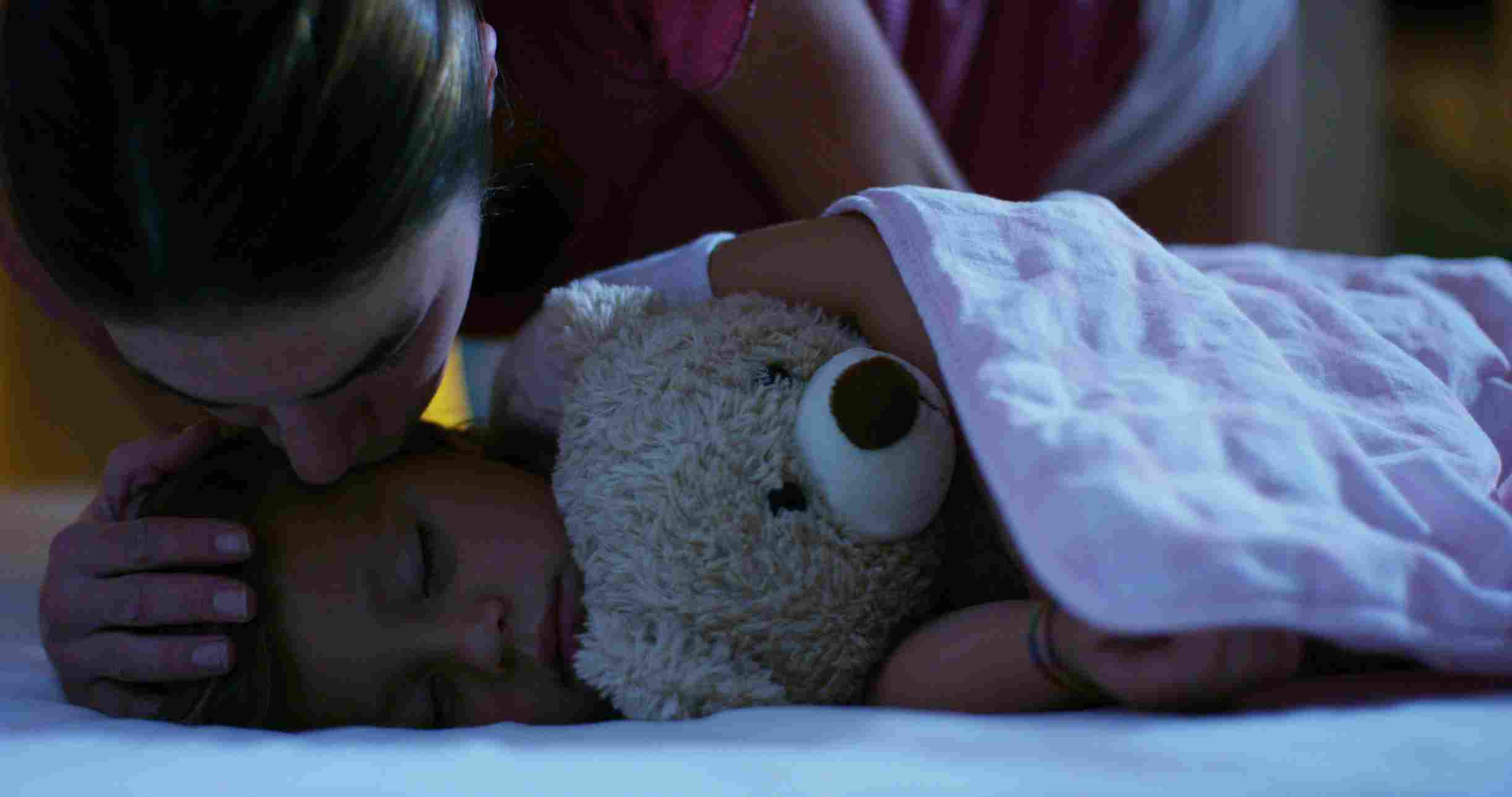 Mama nežno ljubi uspavano dete koje grli plišanog medu tokom večernjeg uspavljivanja