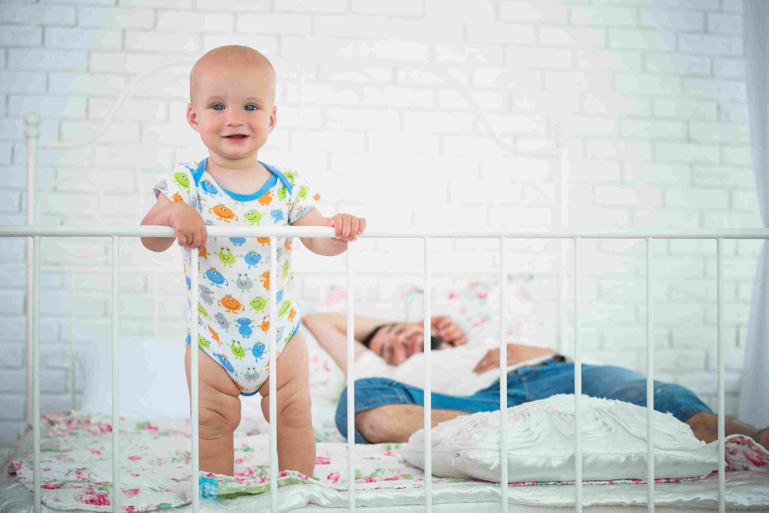 Beba stoji u krevecu dok roditelj odmara u pozadini spavaće sobe.